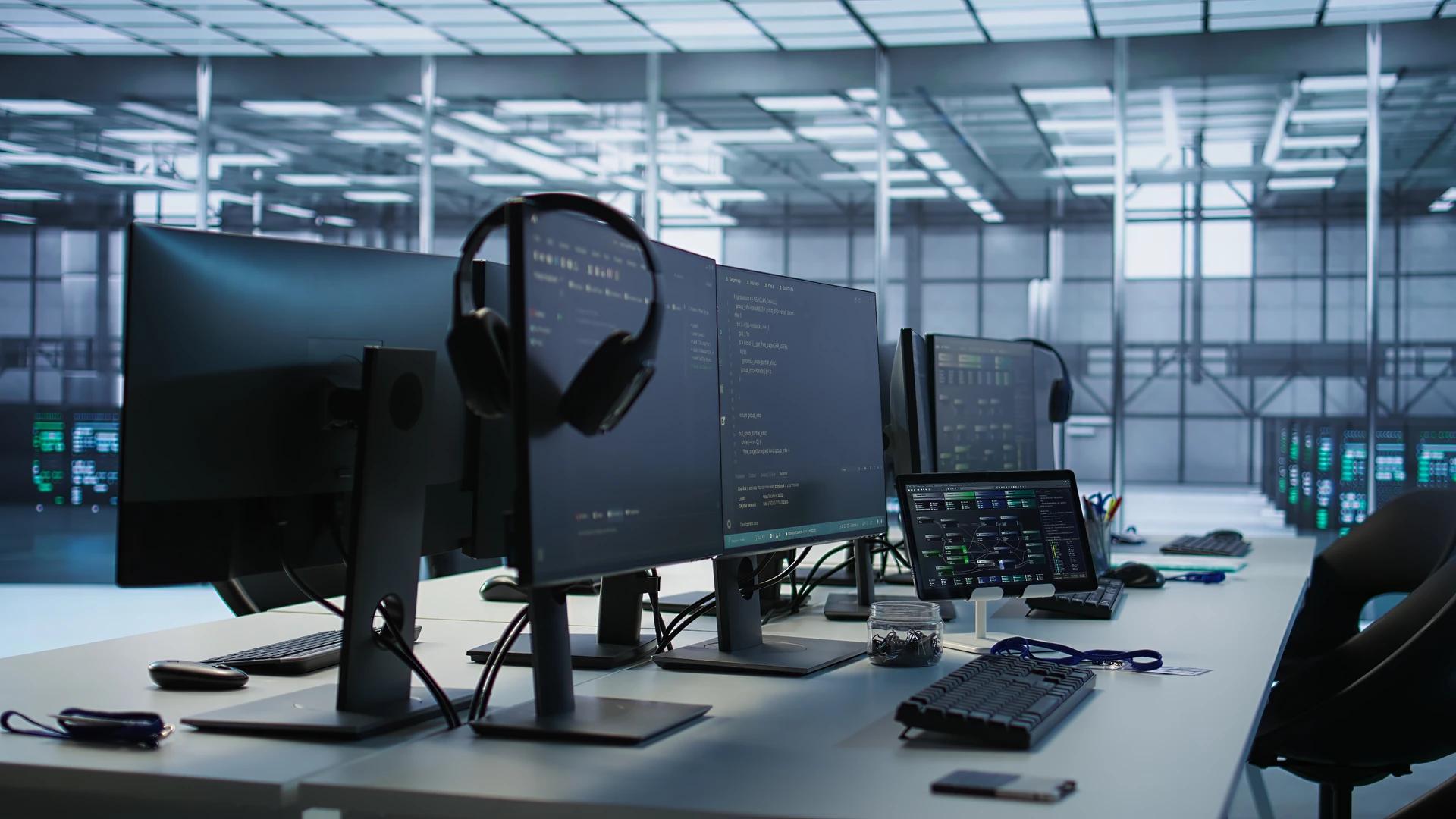 IT workspace with monitors showing code, a tablet with a network diagram, and server racks in the background